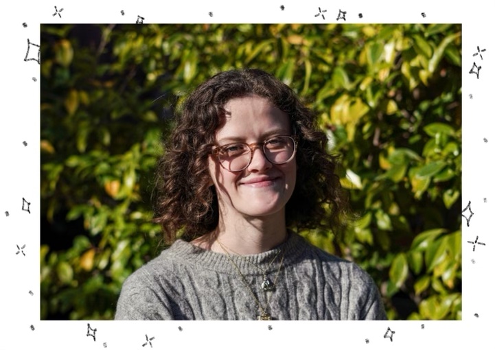 A headshot of Cara Nixon, a woman with curly hair and glasses, in front of greenery at Berkeley Graduate School of Journalism.